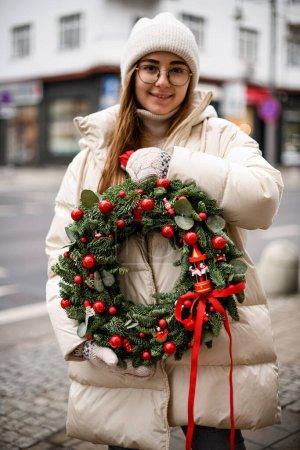 Foto de Joven hermosa mujer sostiene en sus manos corona de Navidad de ramas de abeto y hojas de eucalipto decoradas con bolas y cuentas rojas - Imagen libre de derechos