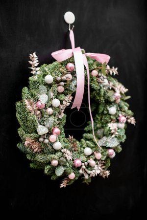 Nahaufnahme auf charmanten Weihnachtskranz aus Tannenzweigen mit rosa Glitzerkugeln und Pflanzen und Schleife auf dunklem Hintergrund