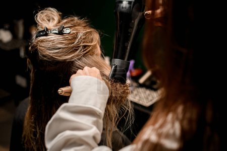 Téléchargez les photos : Female hairdresser stylist skilfully making hairstyle using hair dryer and round comb, blowing on wet customer hair at beauty salon - en image libre de droit