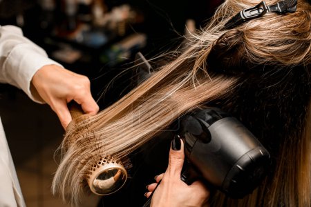 Foto de Close-up of hairdo making process. Hairdresser masterfully puts long female hair with dryer and round comb - Imagen libre de derechos