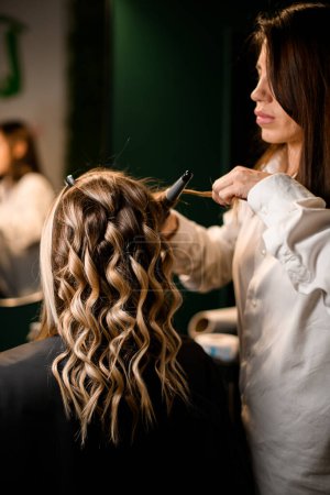 Professional stylist curls long female hair with modern clipless curling iron and makes beautiful hairstyle for woman client.