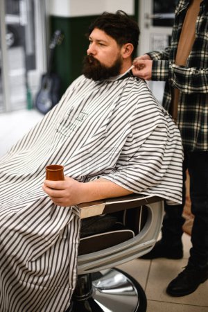 Photo for Man with beard covered with haircut cape sits in hairdressers chair and waits for haircut. Barbershop concept. - Royalty Free Image