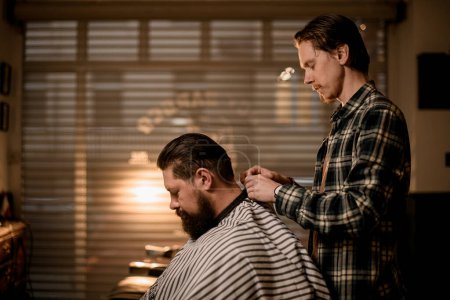 Photo for Side view of bearded male client to whom a male barber puts on hairdresser cape for cutting hair. Barbershop concept - Royalty Free Image