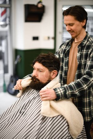 Téléchargez les photos : Man with beard covered with haircut cape sits in hairdressers chair and male barber covers his shoulders with towel. Barbershop concept. - en image libre de droit