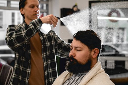 Photo for Male barber splashes from the spray bottle at the head hair of his client with a big beard in the barbershop. - Royalty Free Image