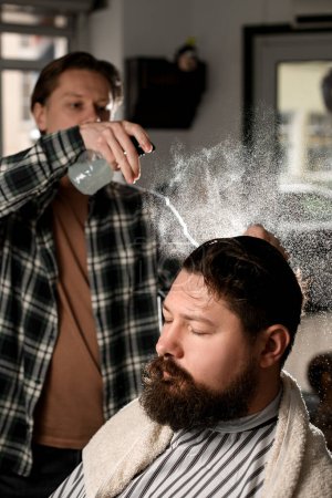 Photo for Male barber gently splashes water from the spray bottle at the head hair of his client with a big beard in the barbershop. - Royalty Free Image