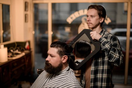 Foto de Side view of bearded man in a barbershop to whom a male barber shows hairstyle with the help of reflection in the mirror - Imagen libre de derechos