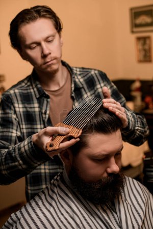 Photo for Male barber doing hair styling to a male client with an afropic comb with long metal teeth - Royalty Free Image