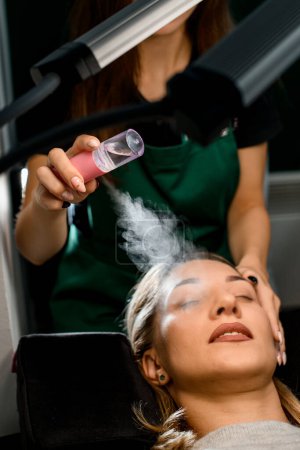 Foto de Gran vista de cerca de cliente femenino y esteticista hidrata su cara con vapor de humidificador portátil. Tratamiento de belleza de la cara en el centro de belleza - Imagen libre de derechos