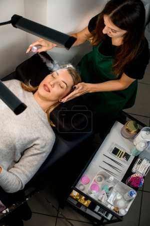 Photo for Top view of female client and beautician carefully moisturizes her face with steam from portable humidifier. Beauty treatment of face in beauty center - Royalty Free Image