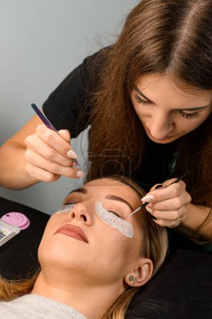 Female cosmetologist with tweezers attaches artificial eyelashes to female client at salon. Eyelash extension procedure.