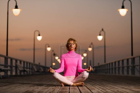 Photo for Front view of woman sitting on wooden quay meditating in lotus pose in solitude on seashore - Royalty Free Image