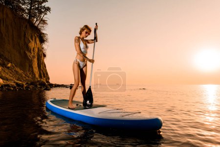 Photo for Woman traveler on stand up paddle board at quiet sea with bright sunset and rock cliff on background - Royalty Free Image