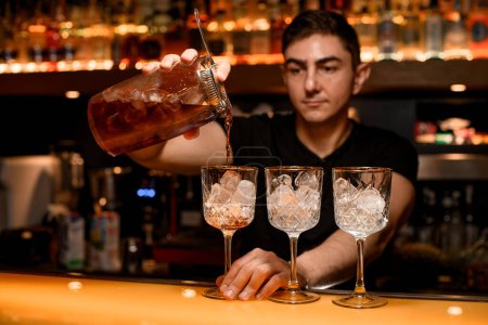 Barkeeper gießt einen Alkohol aus dem Messbecher durch das Sieb in ein Cocktailglas mit Eiswürfeln auf der Theke im unscharfen Hintergrund
