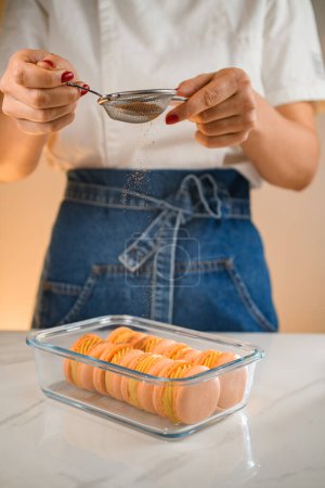 Téléchargez les photos : Femme chef saupoudrer fraîchement fait macarons français avec de la poudre d'orange à l'aide de tamis. Vue rapprochée. Concept de boulangerie maison. - en image libre de droit