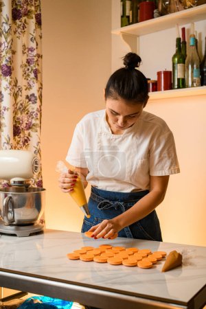 Téléchargez les photos : Femme avec sac de confiserie serrant remplissage de crème aux coquillages de macarons à l'intérieur de la cuisine. Concept de cuisson, de nourriture et de cuisson - en image libre de droit