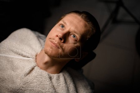 Photo for Handsome young bearded guy with white towel aroun his neck and shoulders sitting in armchair in barber shop. Client waiting for service - Royalty Free Image