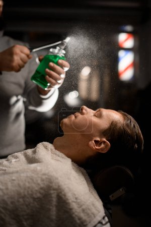 Photo for Professional barber sprinkles green liquid from vintage desidned stylish bottle above clients face in modern barbershop on blurred background - Royalty Free Image