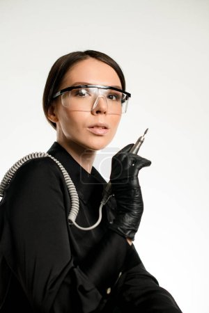 Foto de Manicura femenina en uniforme negro con gafas protectoras en la cara sostiene la máquina de manicura en la mano. Fondo blanco. - Imagen libre de derechos