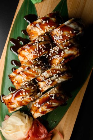 Sushi roll with smoked eel with teriyaki sauce served on wooden board with plant leaves and ginger, top view. black background. Sushi menu.