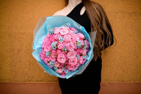 Selective focus on large bouquet of fresh pink roses decorated with small blue flowers formed with blue wrapping paper. Woman with bunch of flowers. Cropped shot