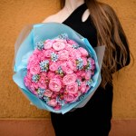 Selective focus on large bouquet of fresh pink roses decorated with small blue flowers formed with blue wrapping paper. Woman with bunch of flowers. Cropped shot