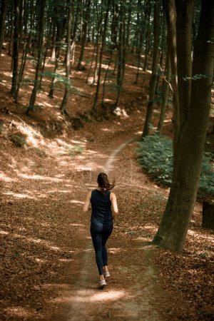 Rückansicht einer fitten Frau, die auf einem schmalen Waldweg zwischen hohen Bäumen läuft. Aktiver Lebensstil. Joggen im Freien.