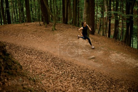 View from above of young woman in black sportswear jogging downhill on forest trail, tall leafy trees on background. Running on fresh air. Healthy lifestyle.