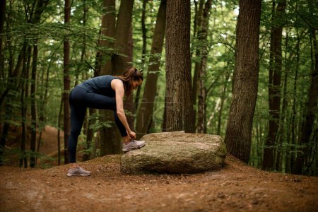 Foto de Corredor mujer se encuentra cerca de piedra grande y atando sus cordones de zapatos, listo para correr a través del bosque.. Estilo de vida saludable activo. Vista lateral - Imagen libre de derechos