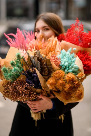 Photo for Young brunette woman florist holding in hands variety of dried decorative flowers on the street. Flower shop. Dried bouquets. Master class and floristry courses. - Royalty Free Image