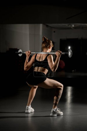 Foto de Vista posterior de la chica muscular en ropa deportiva negro cómodo realizar ejercicios en cuclillas con barra en el gimnasio moderno. Chica entrenando contra el espejo. Estilo de vida deportivo y saludable - Imagen libre de derechos
