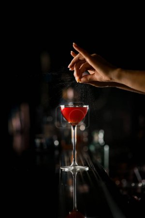 Foto de Mano de camarera femenina espolvorea suavemente jugo de cáscara de naranja sobre vidrio con cóctel alcohólico frío en el mostrador del bar sobre fondo oscuro borroso - Imagen libre de derechos