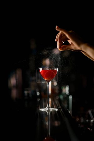 Foto de La mano del camarero espolvorea suavemente jugo de cáscara de naranja sobre vidrio con cóctel alcohólico rojo en el mostrador del bar sobre un fondo oscuro y borroso - Imagen libre de derechos