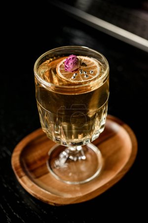 Foto de Vidrio con cóctel transparente decorado con naranja seca, cuentas de plata y pequeña flor de rosa, vista superior. Fondo oscuro - Imagen libre de derechos