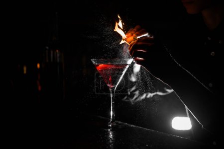 Foto de Copa de cóctel transparente con bebida alcohólica marrón se encuentra en el mostrador del bar, mientras que la mano femenina salpica y sostiene un partido iluminado por encima de ella - Imagen libre de derechos