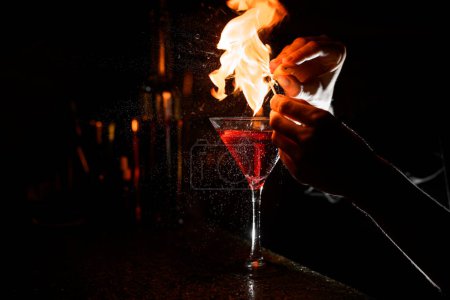 Transparent stemmed cocktail glass with brown alcoholic drink stands on bar counter while female bartender splashes and lights a flame above it in total darkness