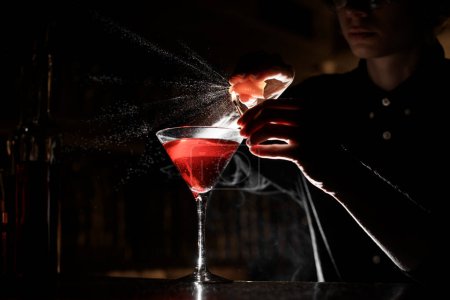 Foto de Barmans manos rociando cáscara de naranja en partido iluminado y la copa de cóctel llena de bebida alcohólica en el fondo oscuro - Imagen libre de derechos