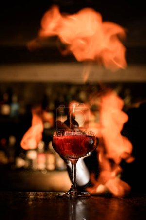 Transparentes Cocktailglas auf Stiel gefüllt mit Cocktail auf Bartheke auf Flammenhintergrund