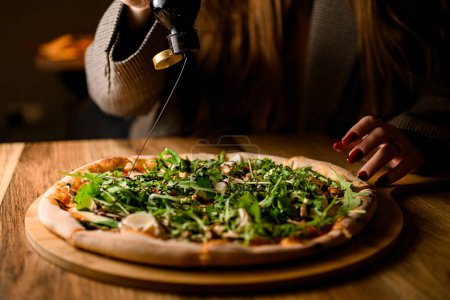 Foto de Momento artístico se desarrolla como una niña delicadamente vierte aceite de oliva sobre una pizza adornada con rúcula, bellamente presentado en una tabla de madera. - Imagen libre de derechos