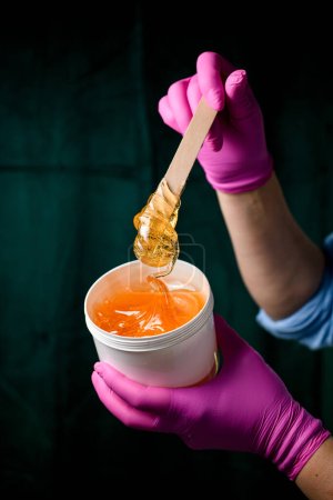 Pasta transparente en un recipiente de plástico en manos del cosmetólogo con una camisa azul y guantes rosados sobre un fondo oscuro