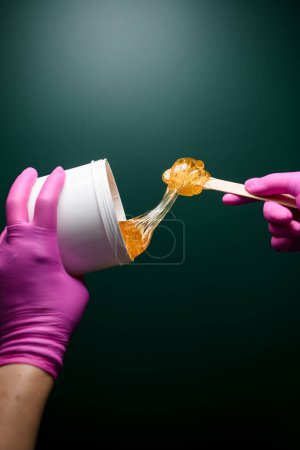 Zuckerpaste in einem weißen Plastikbehälter in einer Hand oder in einem rosa Handschuh auf dunkelgrünem Hintergrund