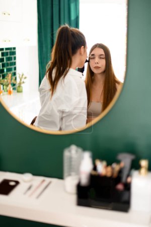 Téléchargez les photos : Dans une image miroir, un styliste en chemise blanche applique de la teinture aux sourcils d'une jolie cliente - en image libre de droit