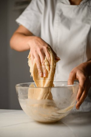 Cook kneads yeast dough with his hands in a deep glass plate. Preparing for further baking in the oven
