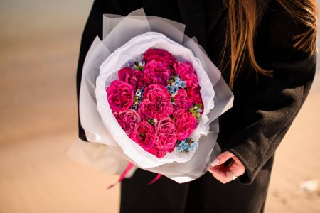 Stylish young brunette woman wearing black coat holding bouquet of bright pink carnations flowers standing by seaside. Top view. Selective focus.