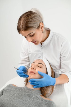 Foto de Cosmetóloga femenina realiza un procedimiento para limpiar la cara de una joven en el área de la barbilla con una cuchara de cosmetología de la ONU, de pie detrás de ella - Imagen libre de derechos