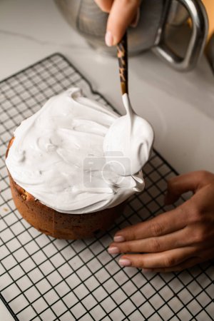 Female hands spread white icing with a spoon on a delicious Easter cake standing on a metal grid