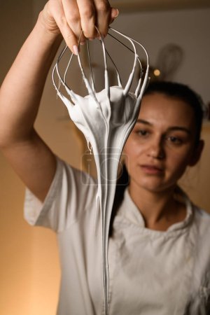 Young brunette woman in white clothes holds a whisk from which white cream flows in the kitchen