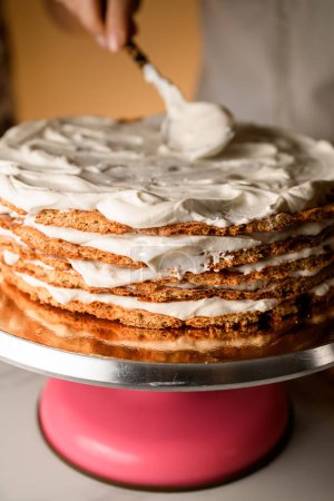 Female hand finishes buttering a round honey cake standing on a pink rotating stand on a white table
