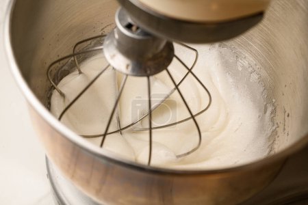 Stand mixer with one large metal whisk whips white cream in a round deep bowl on a white table