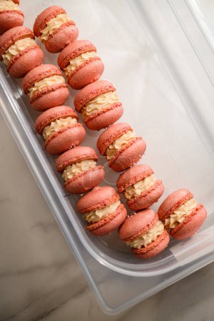 Ready pink beautiful macaroons with white cream neatly lie in a rectangular plastic transparent food container